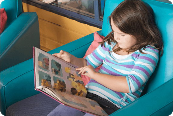 A young girl in a striped shirt sits in a blue vinyl chair. She is flipping a page in a picture book.
