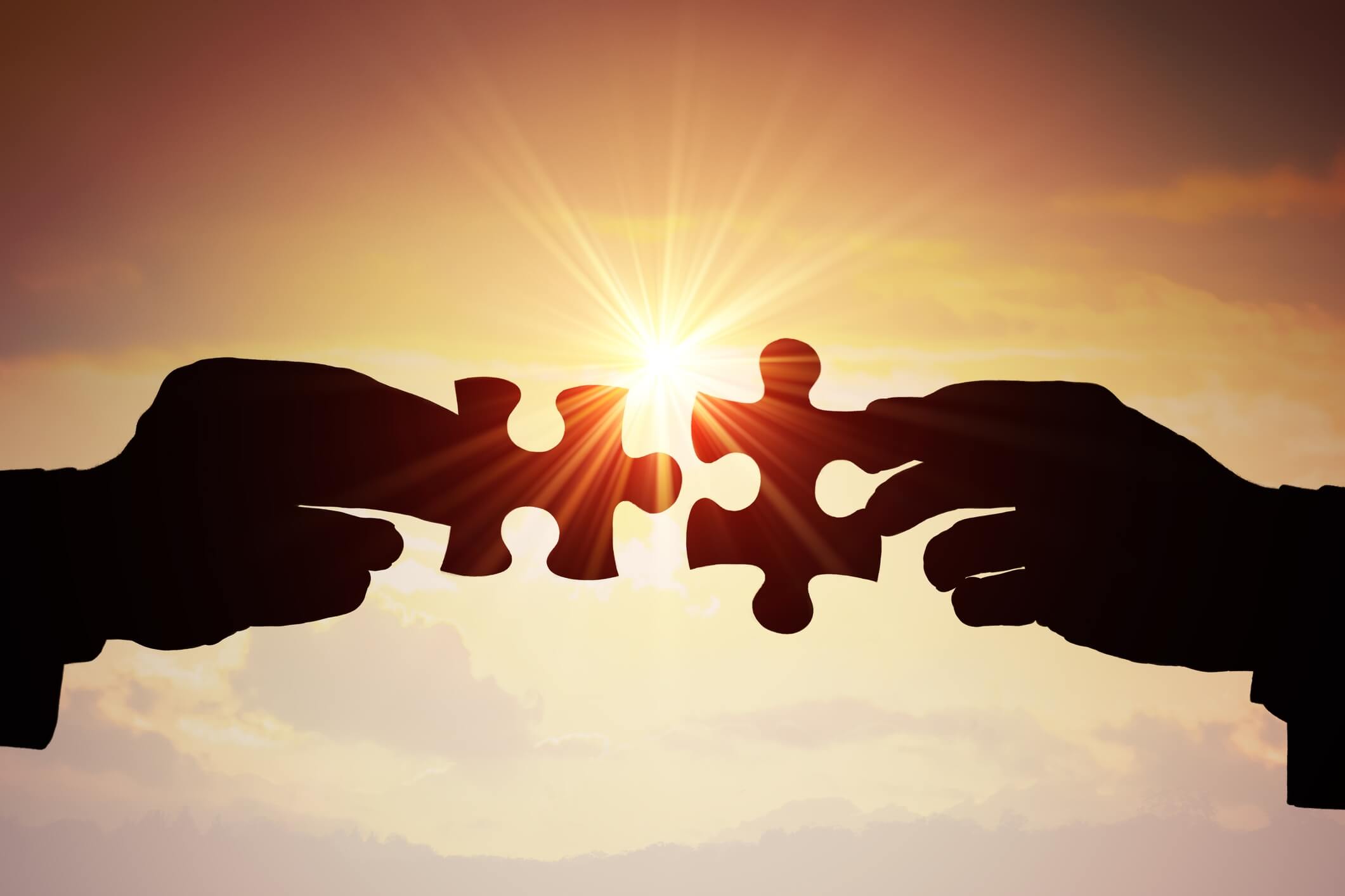 2 hands holding puzzle pieces fitting them together with the sun shining in the background.