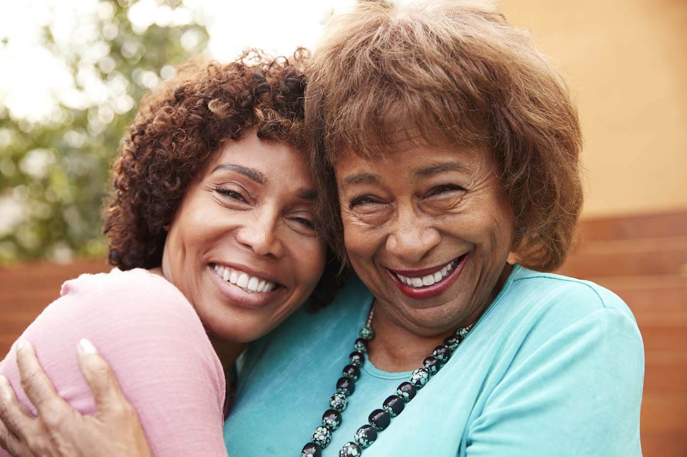 Senior mother in a light blue shirt embraces her middle aged daughter in a pink shirt and smile to camera, close up