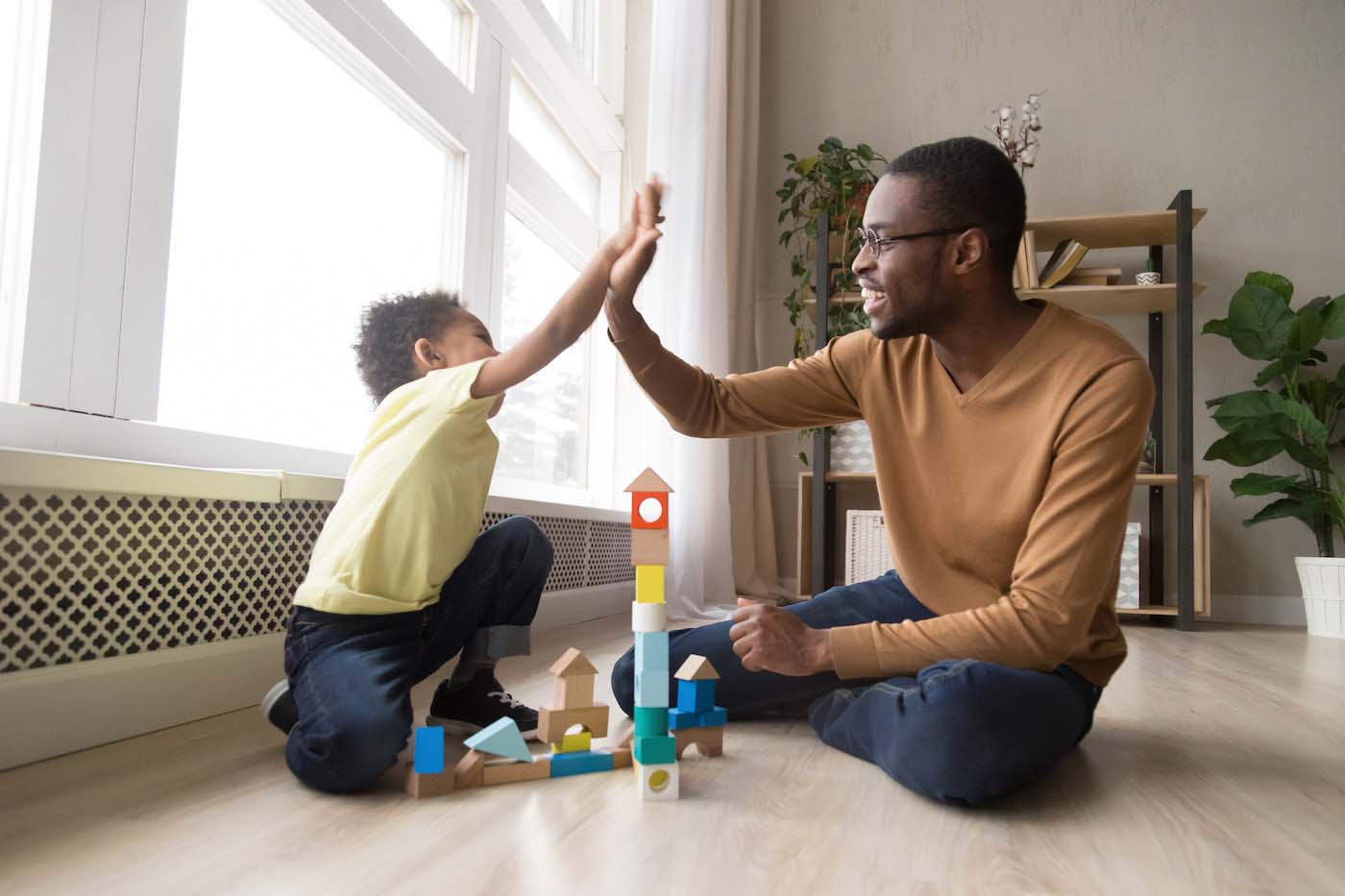 Father and preschooler son sit on the floor and play with building blocks at home. They are high-fiving.