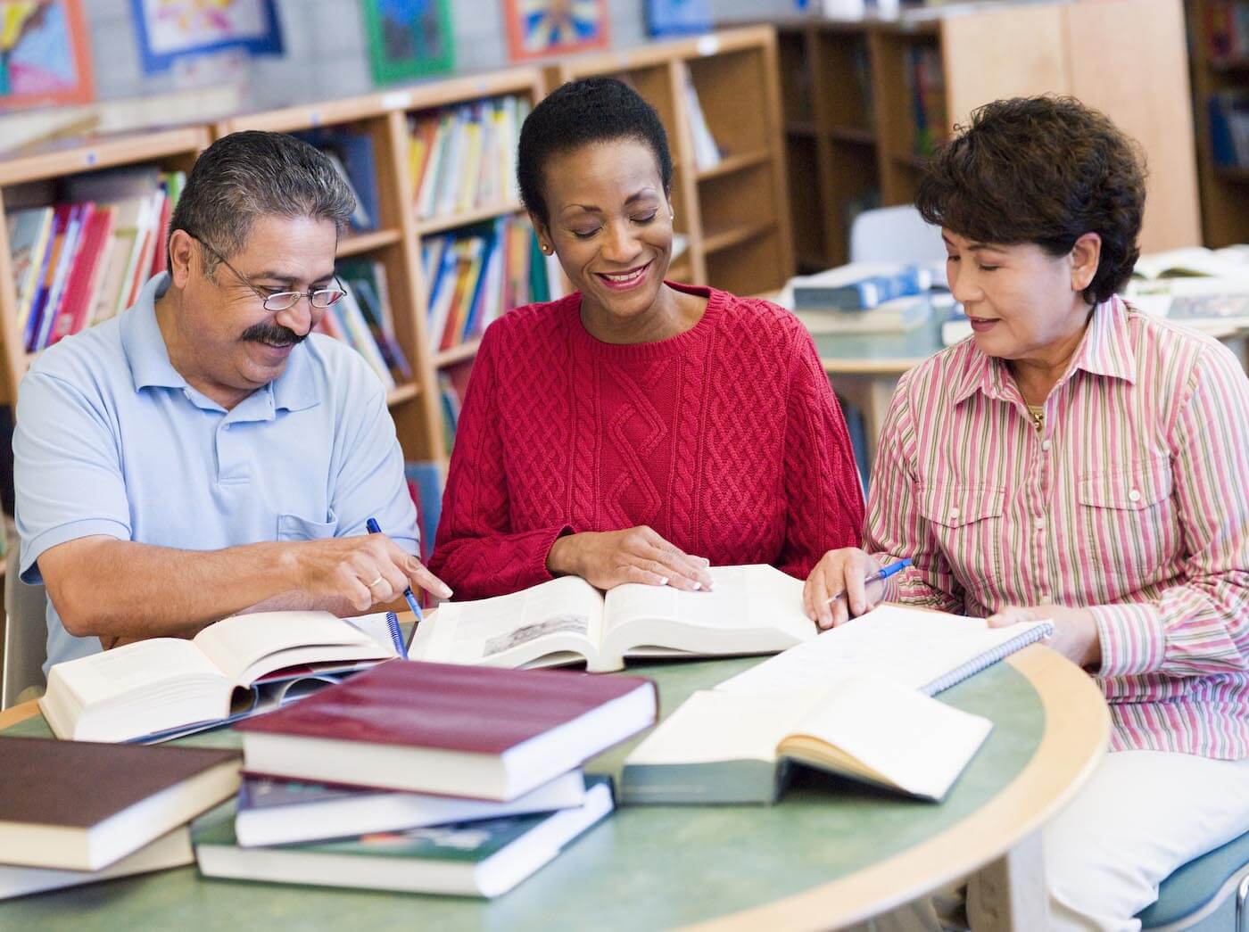 A man and two women sit at a table in a library while looking at their school books and smiling.